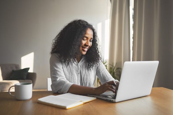 Gorgeous positive young dark skinned female blogger keyboarding on generic laptop, smiling, being inspired while creating new content for her travel blog, sitting at desk with diary and mug